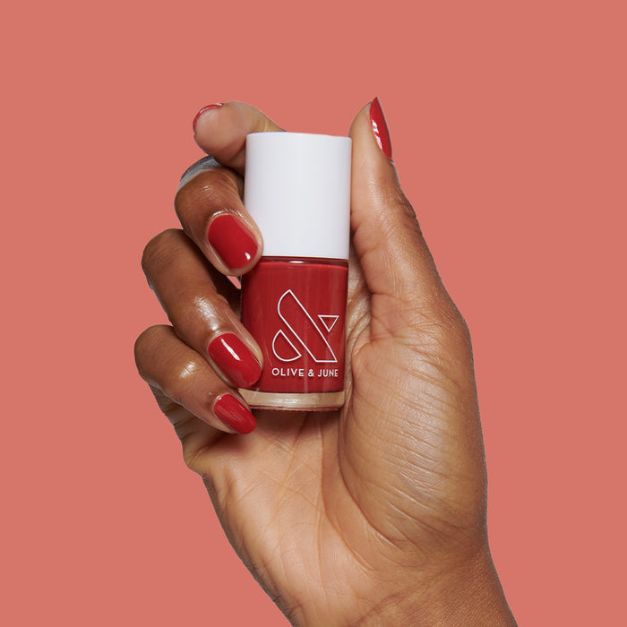 Hand with nails painted in deep berry shade holding Lipstick Stain long-lasting polish bottle against muted red background