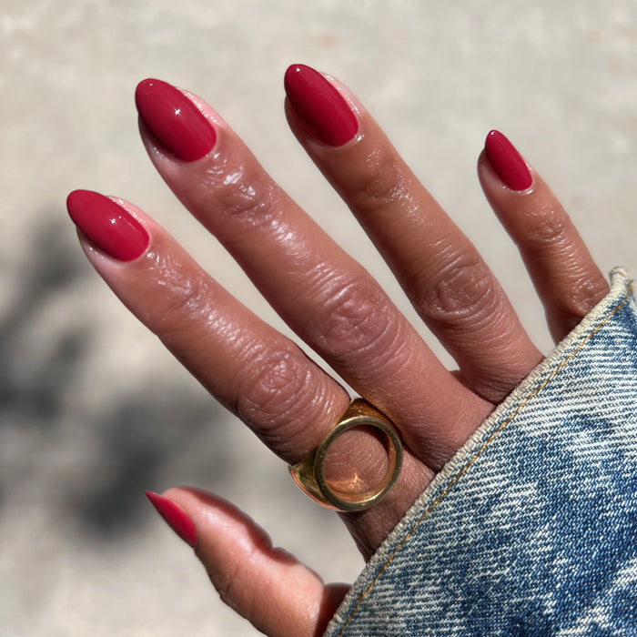 Hand with almond-shaped nails painted with Lipstick Stain deep berry red polish wearing gold ring and denim sleeve outside