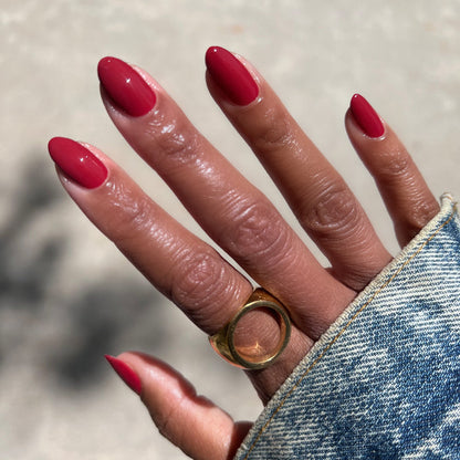 Hand with almond-shaped nails painted with Lipstick Stain deep berry red polish wearing gold ring and denim sleeve outside