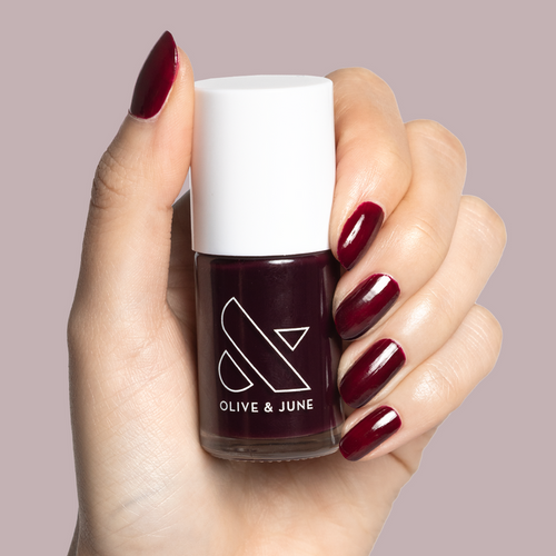 Hand with light skin tone holding Luxe Loafer gel polish bottle with glossy deep red manicure nails visible