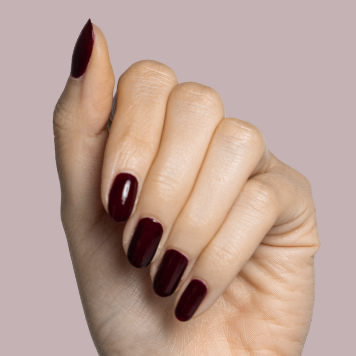 Hand with light skin tone wearing Luxe Loafer gel polish in deep crimson jelly for a glossy manicure on pale mauve background