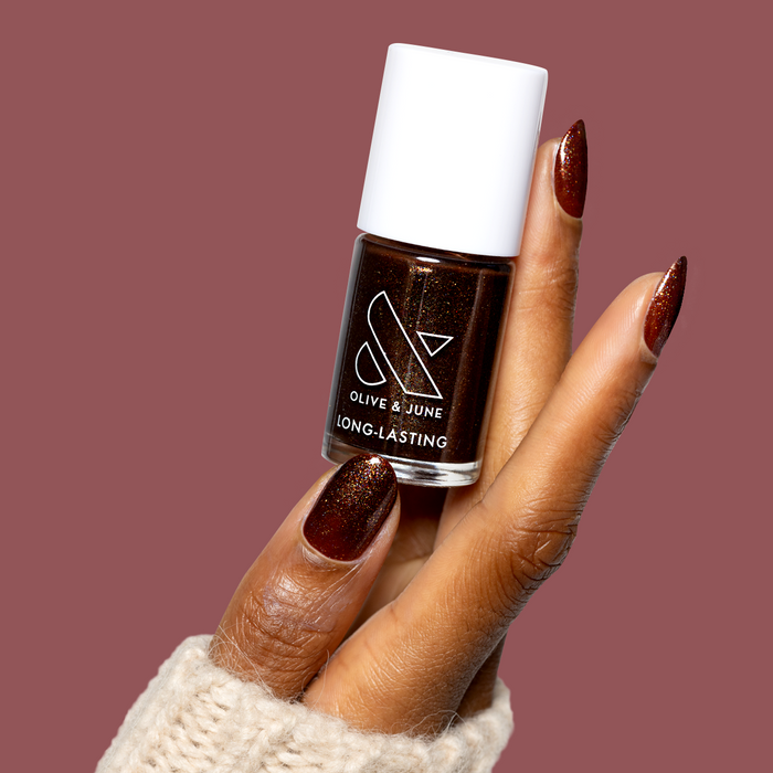 Hand with deep shimmery chocolate brown nails wearing Museum Date gel polish holding Olive & June bottle against mauve background