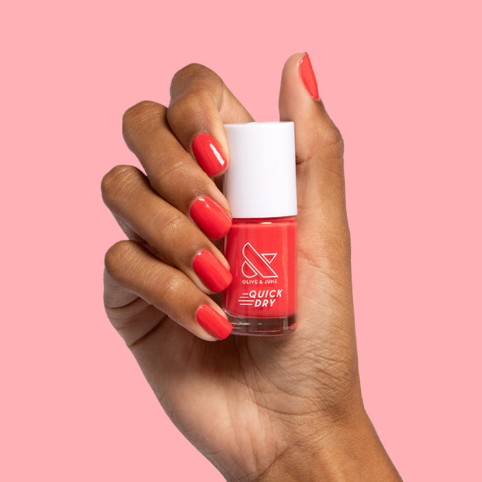 Hand with nails painted in bright orangey red Picante polish holding bottle against soft pink background for mani display