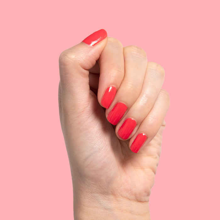 Hand with nails coated in bright orangey red Picante polish against a soft pink background for mani display