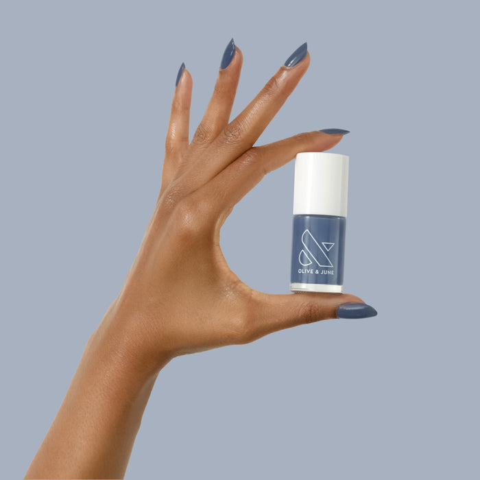 Hand with Social Studies polish in deep sun-bleached navy holding Olive and June bottle against blue background