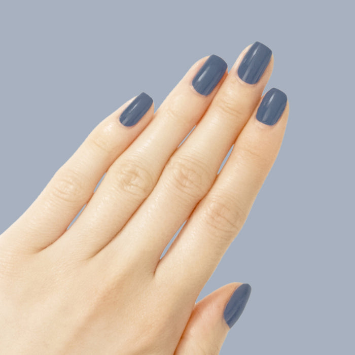 Hand with Social Studies polish in sun-bleached deep navy showing a glossy manicure against a muted blue background