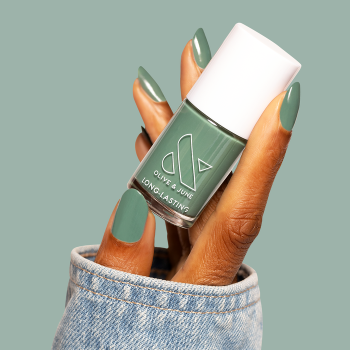 Hand with nails painted Still Life of Flowers holding Olive and June long-lasting gel polish bottle against green background