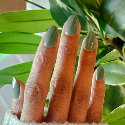 Hand with medium-length nails painted in Still Life of Flowers gel polish in cool mossy green against leafy background