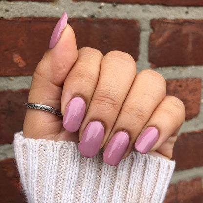 Hand with glossy mauve manicure featuring World Lit polish against a textured brick wall background
